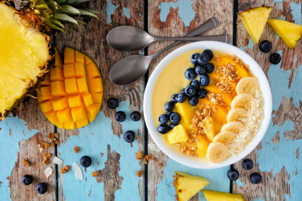 A glass of fresh blueberry pineapple banana smoothie, garnished with blueberries and pineapple chunks, sitting on a wooden table.