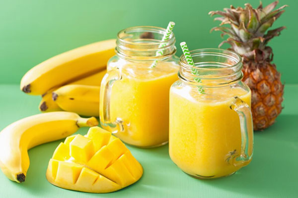 A tall glass of Mango Pineapple Banana Smoothie topped with a slice of pineapple, surrounded by fresh mango, banana, and pineapple chunks.