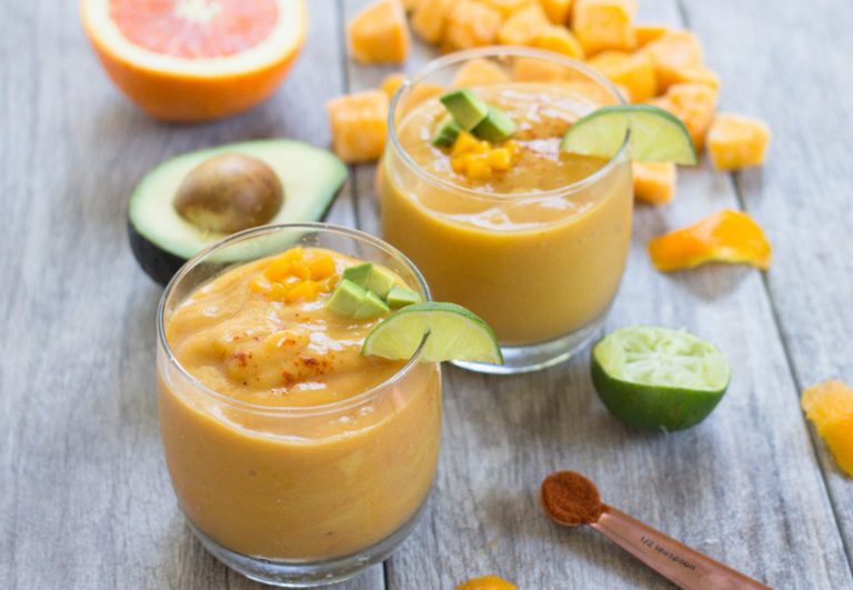 A refreshing glass of creamy Avocado Mango Orange Smoothie, garnished with a wedge of lime, surrounded by fresh mango chunks, a halved avocado, and a halved orange on a wooden table.