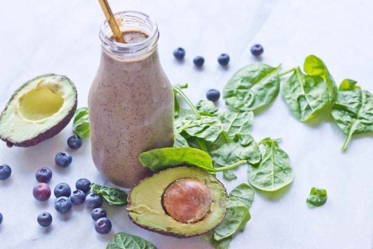 A glass of vibrant green Blueberry Avocado Spinach Smoothie topped with fresh blueberries and a sprig of spinach, sitting on a wooden table.