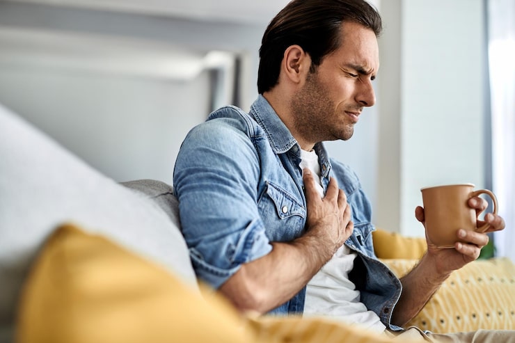 A young man experiencing chest pain at home, holding his chest while drinking tea — early warning sign of arterial stiffness, high blood pressure, and vascular aging. Learn how to reverse arterial stiffness at any age.