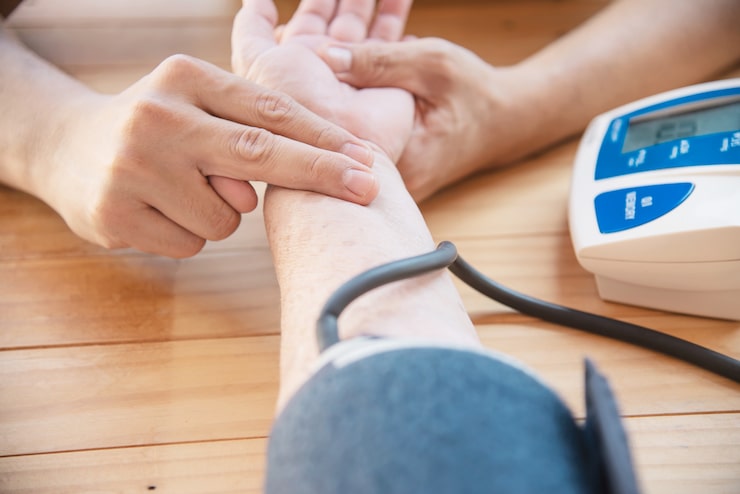 Healthcare provider checking a patient's pulse while a blood pressure monitor sits nearby — illustrating how pulse pressure is measured and monitored for cardiovascular health assessment. Read more on widened pulse pressure.