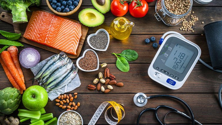 Top 10 foods that lower blood pressure including salmon, sardines, pistachios, blueberries, avocado, spinach, olive oil, carrots, and a blood pressure monitor on a wooden table.