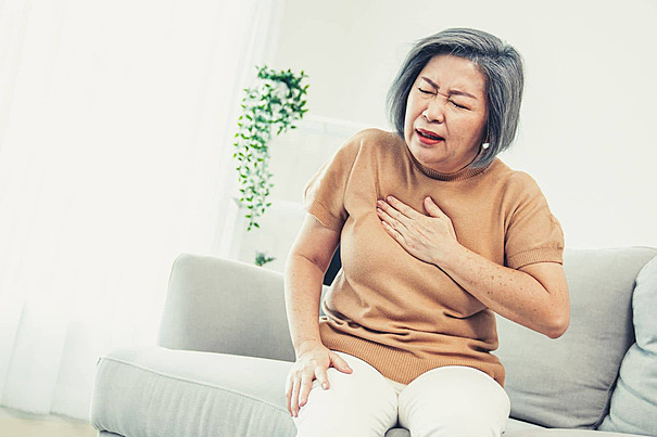 Older woman clutching her chest in pain on a couch, showing signs of a heart attack caused by coronary thrombosis. Coronary thrombosis vs Embolism: Learn the angiographic, treatment, and diagnostic differences backed by research.